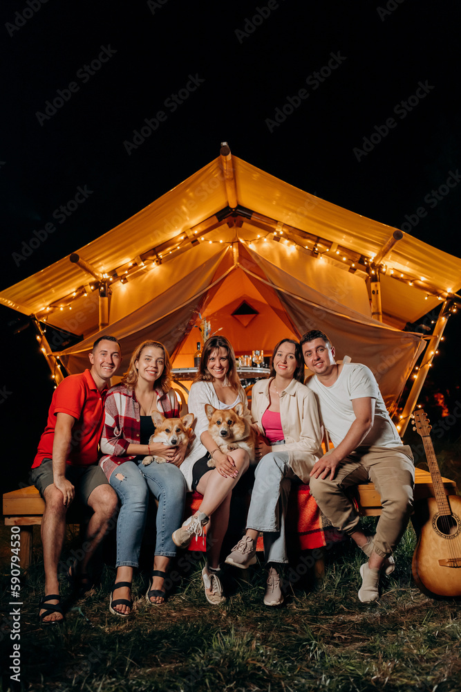 Happy group of friends with Welsh Corgi Pembroke dogs relaxing in glamping on summer evening near cozy bonfire. Luxury camping tent for outdoor recreation and recreation. Lifestyle concept