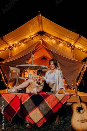 Happy young woman with her Welsh Corgi Pembroke dog relaxing in glamping on summer evening near cozy bonfire. Luxury camping tent for outdoor recreation and recreation. Lifestyle concept