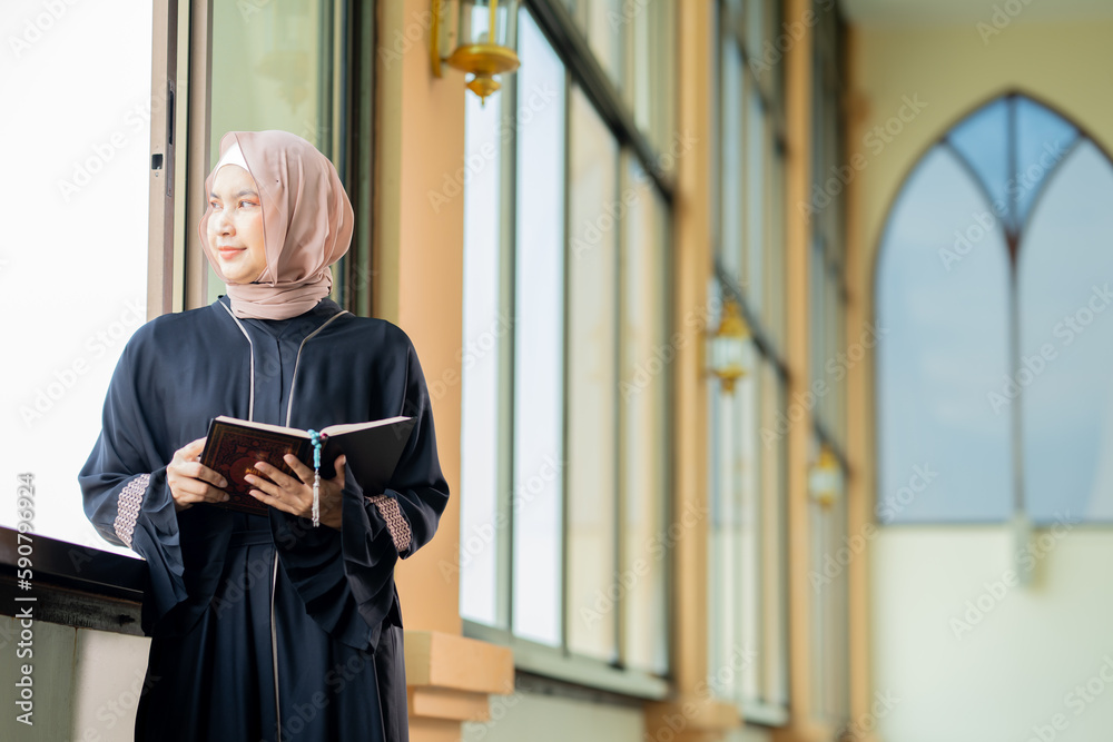 The image of an Asian Muslim woman in the Islamic religion in hijab in cream color. reading the Quran and having a happy smiling face Staying in a beautiful mosque out of respect for God.