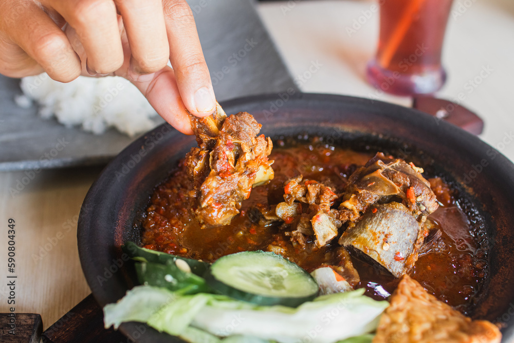 Fototapeta premium Sambal iga or ribs with sambal cooked in earthenware plate against wooden background. Served on wooden table