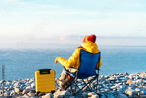 Woman in yellow coat and orange scarf reaching the destination and sitting on beach chairs on the seaside at sunrise. British cold winter. Local tourism concep © Iryna