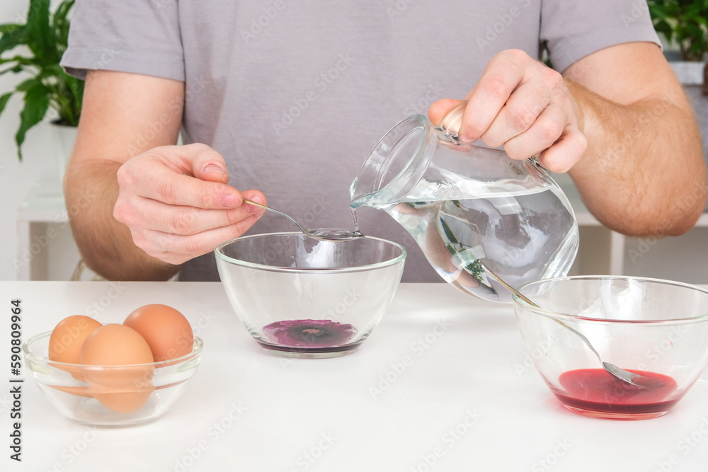A man pours spoonful water into glass cup with dye. Dyeing eggs with coloring in Easter egg A man pours spoonful water into glass cup with dye. Dyeing eggs with coloring in Easter egg