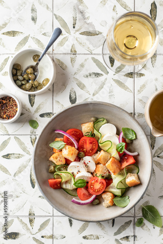 Traditional italian tomato salad panzanella with mozzarella, capers, red onion, croutons, cucumbers and basil with fork and wine glass. Summer salad on printed tile background top view