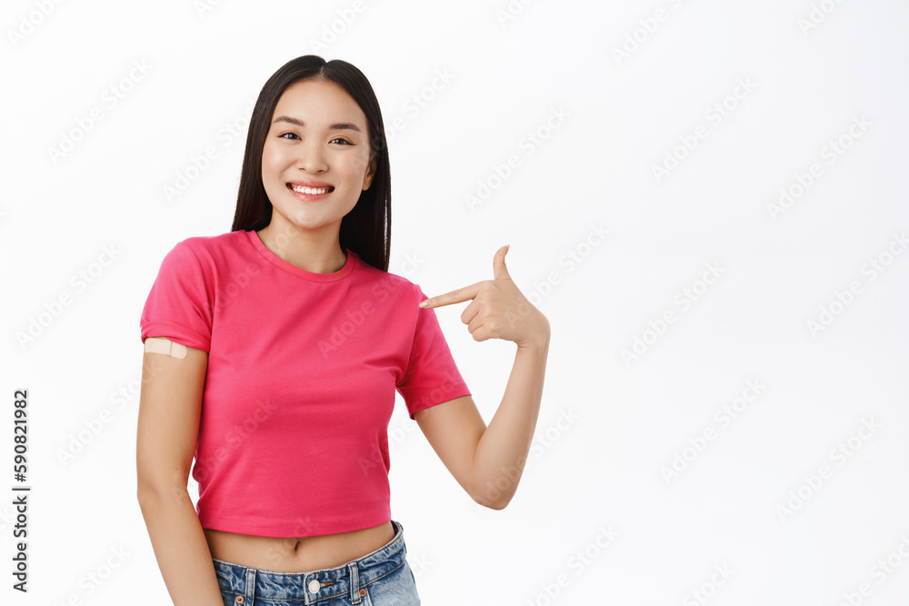 She got covid-19 vaccine. Smiling asian girl points at her shoulder with band aid from coronavirus vaccination, standing over white background She got covid-19 vaccine. Smiling asian girl points at her shoulder with band aid from coronavirus vaccination, standing over white background