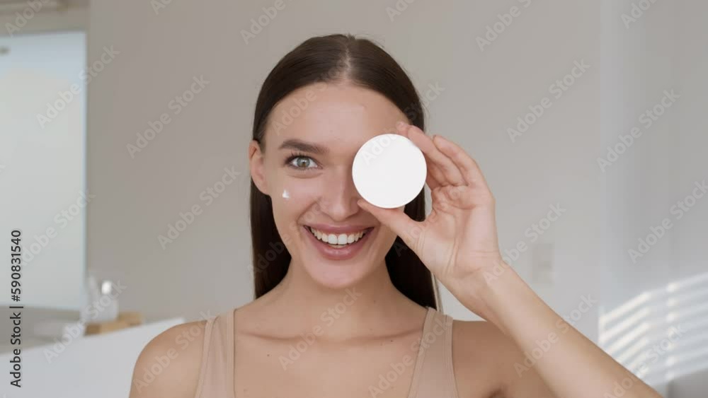 Facial skincare concept. Young happy beautiful woman with cream spots on cheeks laughing to camera, holding jar near eye Facial skincare concept. Young happy beautiful woman with cream spots on cheeks laughing to camera, holding jar near eye