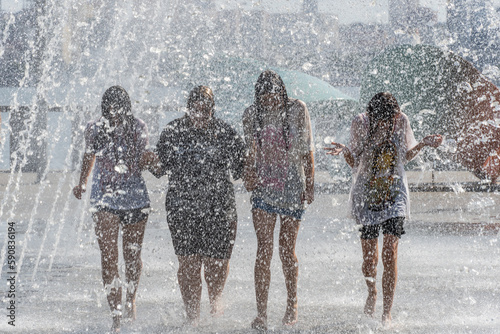 Group sopping girls splashing under jets in pedestrian fountain on hot sunny day. Young womans enjoy cool water in heat summertime. Happy youth fun in city floor archwise fountain. Persons play drops.