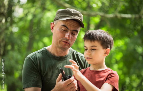 Military man in olive uniform and cap showing his little son radio set outdoors in park © Olena Shvets