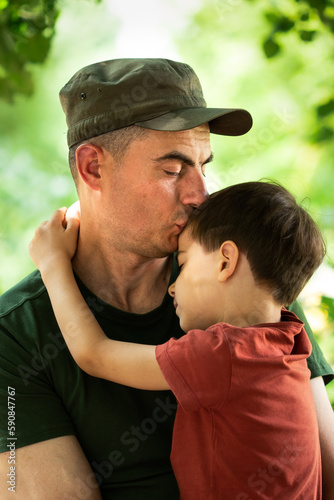 Military man in olive uniform and cap hugging and kissing his little son and smiling outdoors in park © Olena Shvets