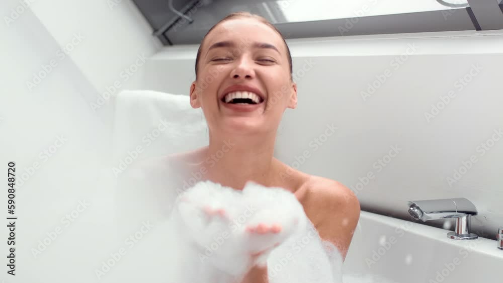 Close up portrait of young happy woman having hot bath at home, blowing soap foam to camera and laughing, slow motion