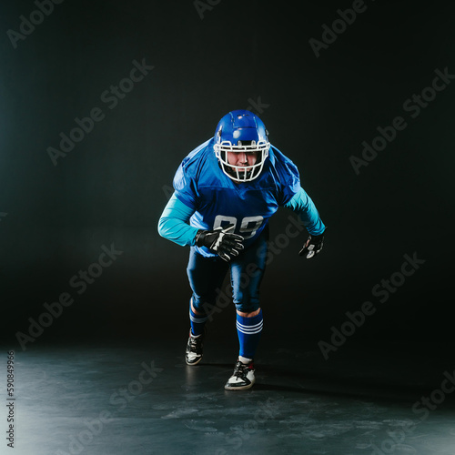 Portrait of a man in a blue uniform for american football runs with a ball on a black background. 