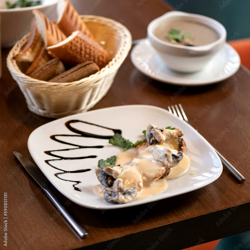 Champignon mushrooms in creamy sauce and champignon cream soup. Lunch at restaurant. Two dishes, basket of bread, fork and knife on table. Wooden table. Side view. Soft focus.