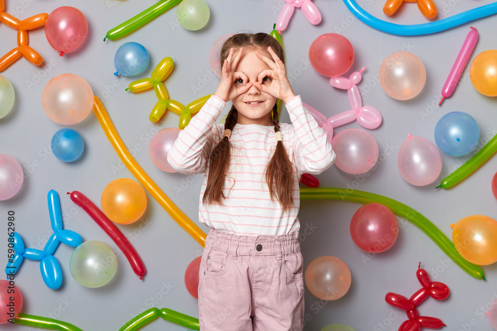 Fototapeta premium Funny smiling little girl with pigtails standing against gray wall decorated with colorful balloons making binocular gesture looking far waiting for guests having birthday party.