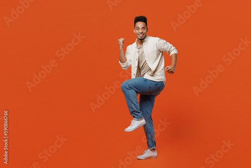 Full body overjoyed young man of African American ethnicity wear light shirt casual clothes doing winner gesture celebrate clenching fists say yes isolated on orange red background studio portrait..