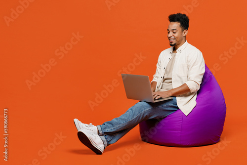 Full body young IT man of African American ethnicity wear light shirt casual clothes sit in bag chair hold use work on laptop pc computer isolated on orange red background studio. Lifestyle concept.