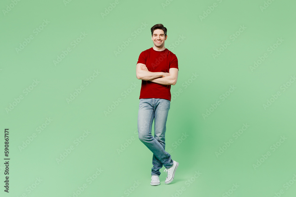 Full body smiling happy fun young brunet caucasian man he wears red t-shirt casual clothes hold hands crossed folded isolated on plain pastel light green background studio portrait. Lifestyle concept.