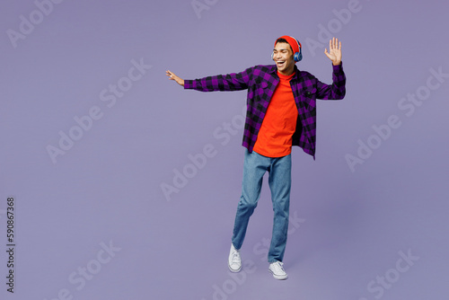 Full body young man of African American ethnicity wear casual shirt orange hat headphones listen to music dance raise up hands isolated on plain pastel light purple color background studio portrait.