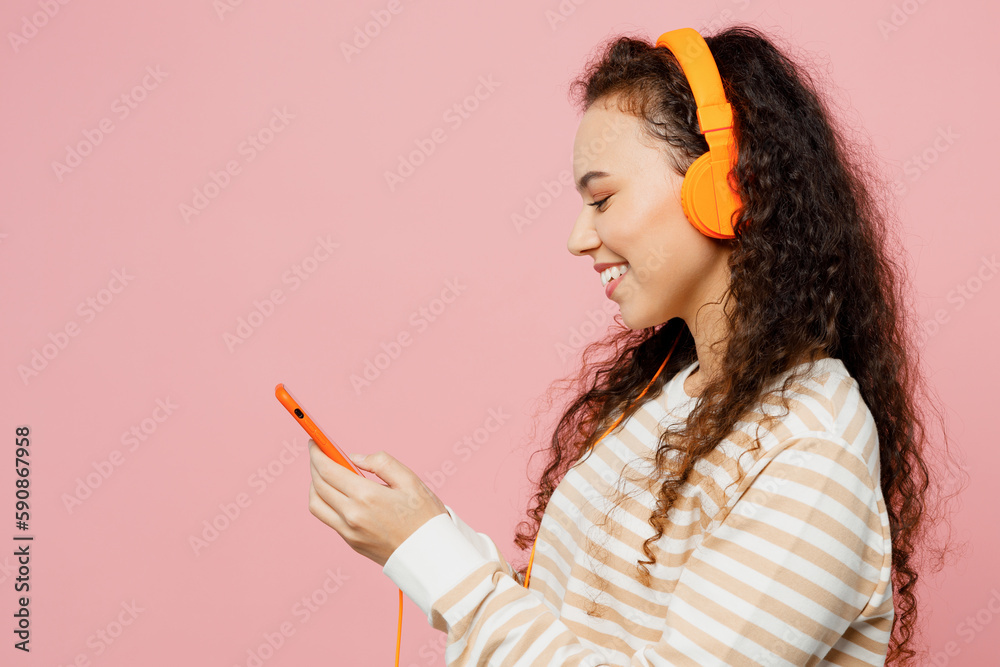 Side view young cheerful fun cool woman of African American ethnicity she wearing light casual clothes headphones listen to music use mobile cell phone isolated on plain pastel pink background studio. Side view young cheerful fun cool woman of African American ethnicity she wearing light casual clothes headphones listen to music use mobile cell phone isolated on plain pastel pink background studio.