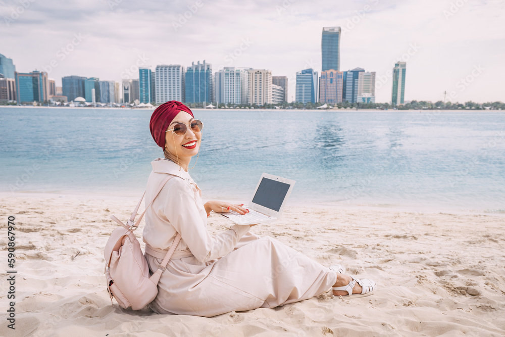 With a laptop open, a young asian woman works remotely on the beach, taking advantage of the stunning view of Abu Dhabi's skyscrapers as her backdrop.