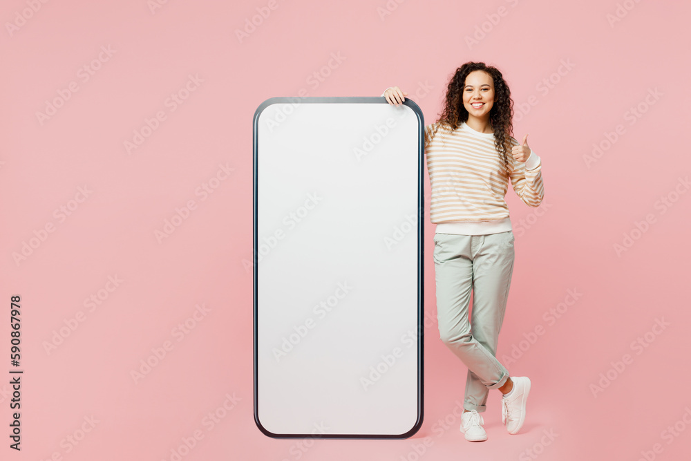 Full body happy young woman wear light casual clothes big huge blank screen mobile cell phone smartphone with area show thumb up look camera isolated on plain pastel pink background studio portrait. Full body happy young woman wear light casual clothes big huge blank screen mobile cell phone smartphone with area show thumb up look camera isolated on plain pastel pink background studio portrait.