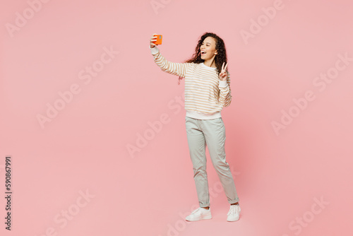 Full body young woman of African American ethnicity wear light casual clothes doing selfie shot on mobile cell phone post photo on social network show v-sign isolated on plain pastel pink background.