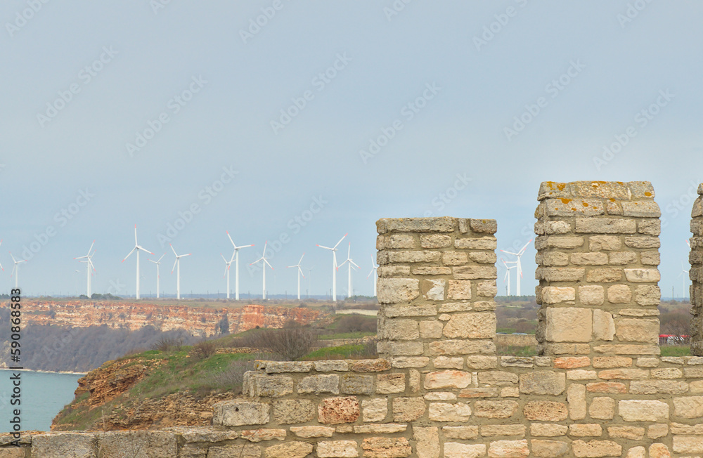 In the foreground, the walls of an ancient fortress in contrast against the background of modern fields are wind generators on the coast of the sea.