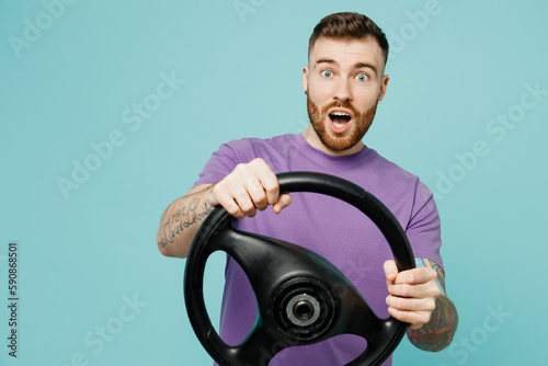 Young shocked sad man wear purple t-shirt hold steering wheel driving car look aside on area isolated on plain pastel light blue cyan color background studio portrait. Tattoo translates life is fight.