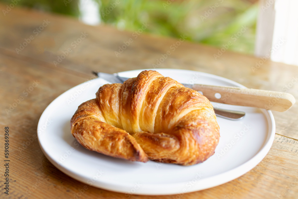 French croissant on white plate on wooden table and nature sunlight with shadow through from window.