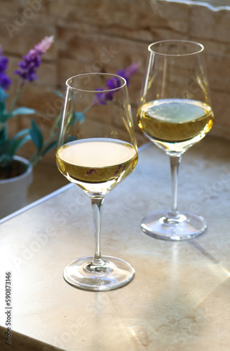 Two glasses of white wine on the kitchen table in front of the window with a purple flower on the back on a sunny day. Close up of a shining glasses of white wine on a table in natural lights