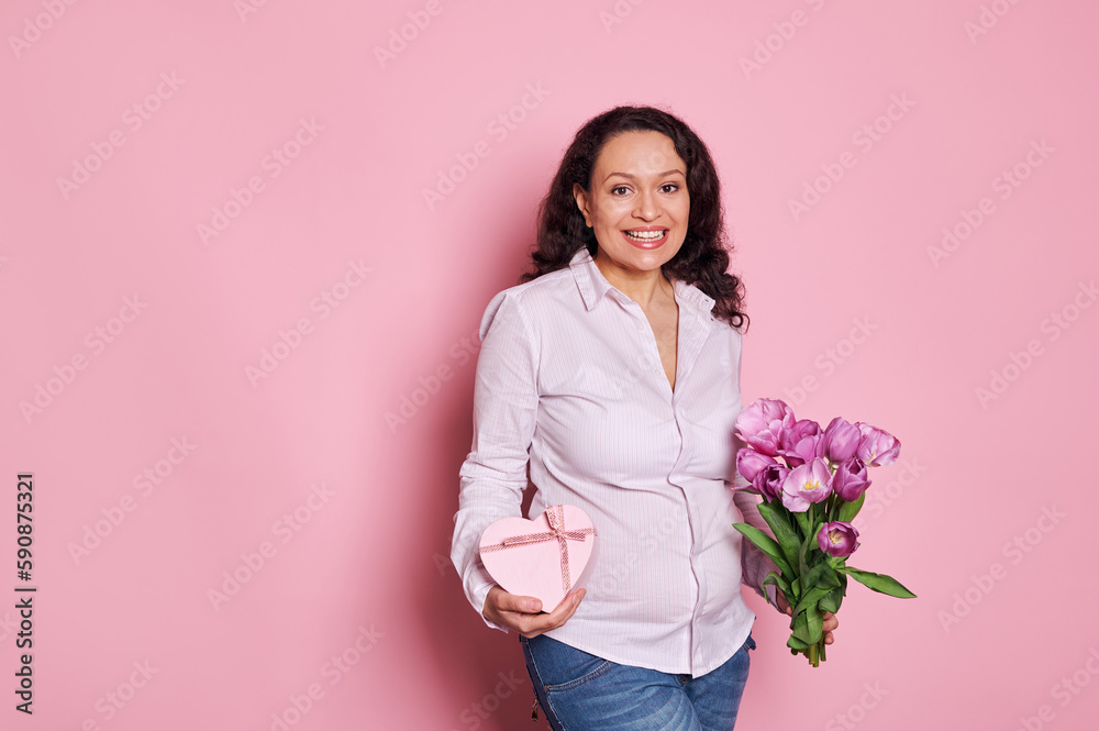 Attractive positive Latin American pregnant woman smiles looking at camera, expresses happiness getting happy present in heart shaped gift box and bouquet of tulips for Mother's Day, isolated on pink Attractive positive Latin American pregnant woman smiles looking at camera, expresses happiness getting happy present in heart shaped gift box and bouquet of tulips for Mother's Day, isolated on pink
