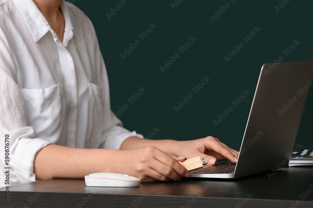 Woman using USB flash drive with modern laptop at dark table near color wall