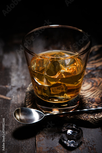 Glass of single malt whiskey with bar spoon and ice cubes on wooden background.