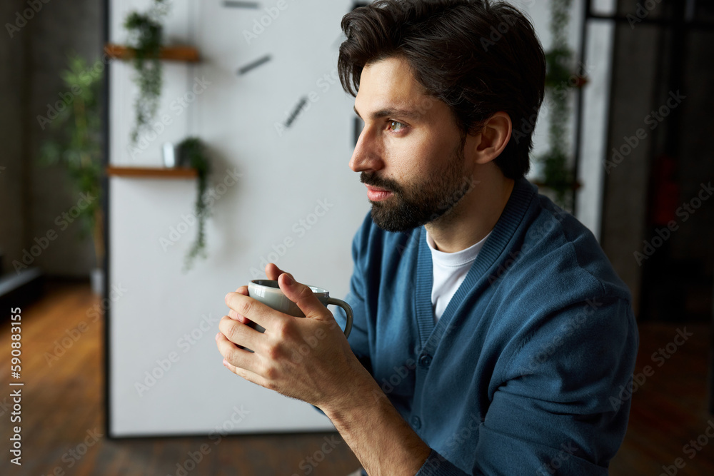 Profile view portrait of good-looking young caucasian guy in blue cardigan holding hot drink in hands, trying to warm himself up and relax, looking at something with pensive facial expression Profile view portrait of good-looking young caucasian guy in blue cardigan holding hot drink in hands, trying to warm himself up and relax, looking at something with pensive facial expression