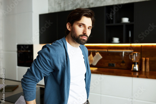 Indoor image of attractive bearded guy freelance worker in stylish cardigan standing against kitchen background leaning on table with his working place and documents, looking at camera