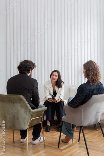 A female psychologist sitting opposite of family couple with a notebook in her hands and attentively listens to her clients at the reception during a psychological counseling