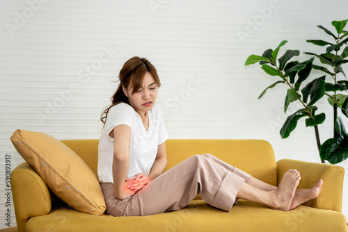 Bild auf Leinwand An Asian attractive woman sitting on the sofa, she is sick and  stomachache or Period cramps, concept to Disease of every woman and health care concept