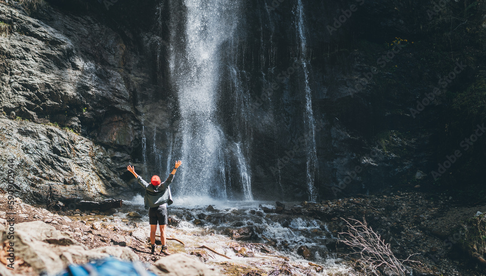 Φωτογραφία A woman dressed in active trekking clothes standing near mountain river waterfall rose arms up and enjoying the splashing Nature power