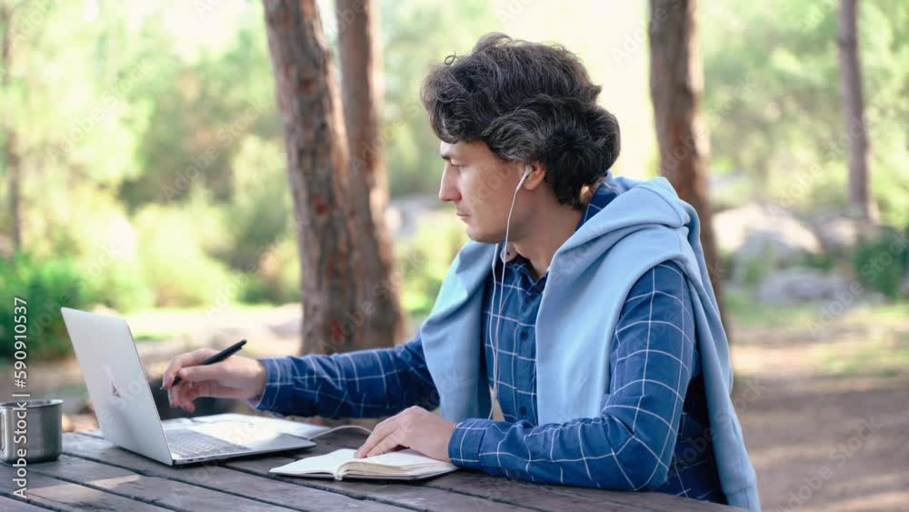 Happy cheerful man with a laptop sitting outdoors in nature forest park. Freelancer with computer writing at notepad in vacation in mountains. Freelance, distance work concept. Happy cheerful man with a laptop sitting outdoors in nature forest park. Freelancer with computer writing at notepad in vacation in mountains. Freelance, distance work concept.