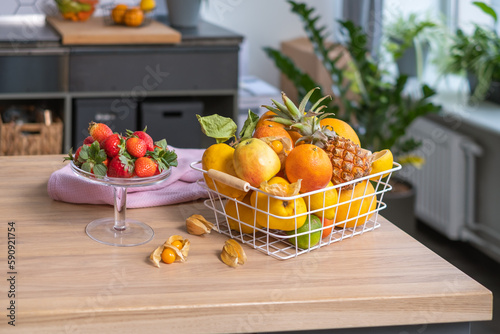 Fototapeta Naklejka Na Ścianę i Meble -  Juicy fruits in a metal basket, strawberries in a glass bowl, physalis and a linen napkin on a light wooden table. A shelf and green houseplants are in the blurred background.