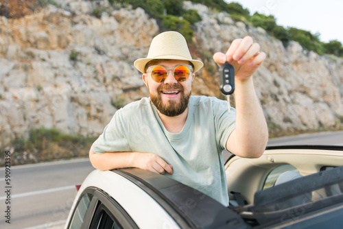 Car driver man smiling showing new car keys and car. Bearded guy driving rented cabrio on summer vacation