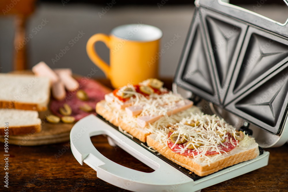 Cooking sandwiches with cheese, sausage, olives in a sandwich maker on a wooden background. Cooking sandwiches with cheese, sausage, olives in a sandwich maker on a wooden background.