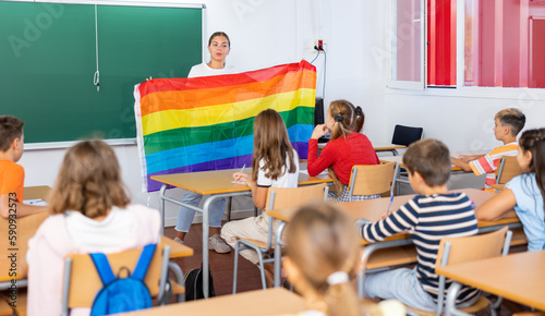 Young woman teacher holding rainbow flag while explaining lesson about LGBTQ people to young girls and boys.