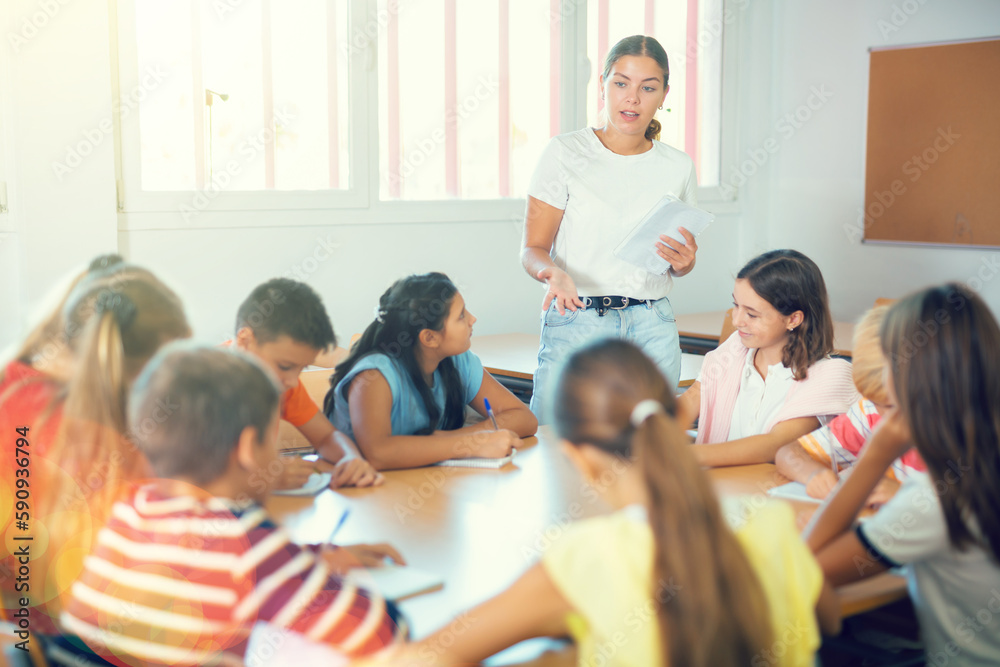 Obraz premium Young female teacher conducting lesson with interested preteen children sitting around common desk in classroom