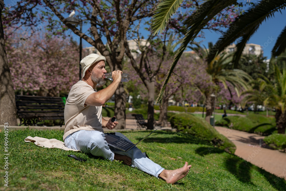 Fototapeta premium bearded male freelancer in the park with a laptop drinks water from a bottle while sitting on the grass with bare feet