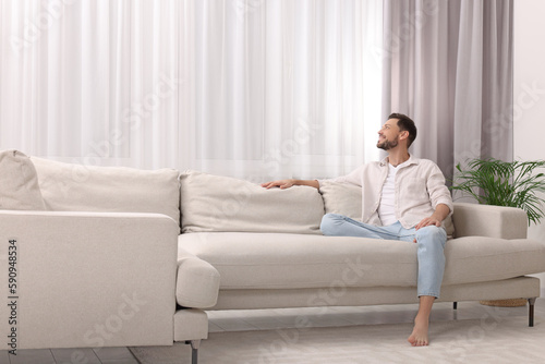 Happy man on resting sofa near window with beautiful curtains in living room. Space for text