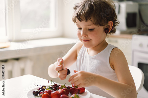 Cute beautiful little boy eating fresh cherry and strawberry. Healthy food, childhood and development. Happy kid at home.