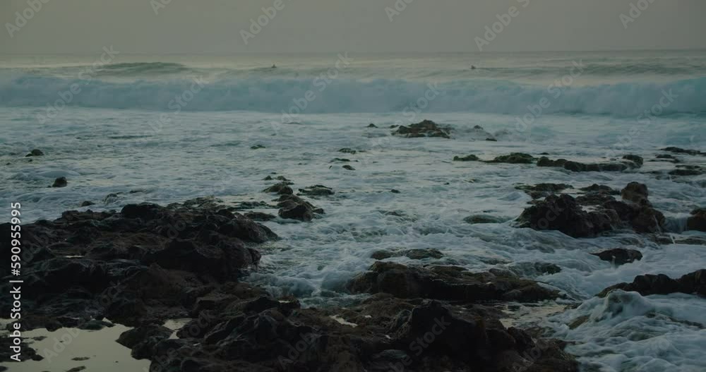 Ocean stormy waves roll on rocky beach. White sea foam splash crash on stones. Moody dark sky at dusk. Surfers wsim in water. Ocean stormy waves roll on rocky beach. White sea foam splash crash on stones. Moody dark sky at dusk. Surfers wsim in water.
