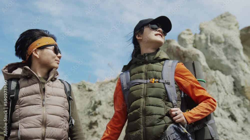 Medium shot of two Asian female friends, tourists with rucksacks, holding hands standing at base of rocks, looking around, smiling, enjoying themselves, wind blowing their hair Medium shot of two Asian female friends, tourists with rucksacks, holding hands standing at base of rocks, looking around, smiling, enjoying themselves, wind blowing their hair