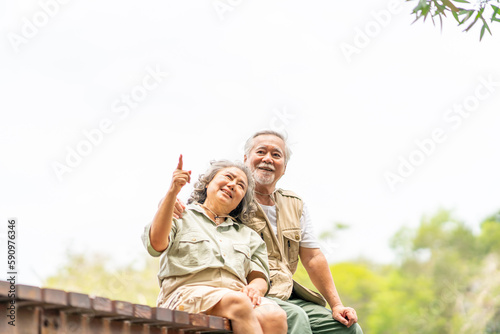 Happy Asian senior couple resting on wooden bridge and looking beautiful nature at tropical forest. Retired elderly people enjoy outdoor lifestyle travel nature hiking together on summer vacation.