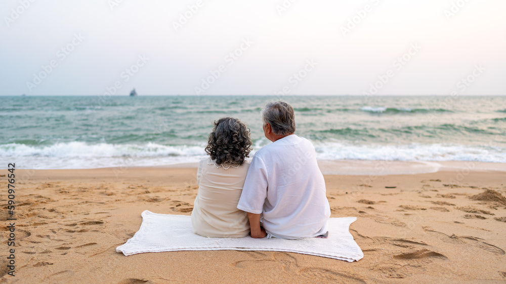 Happy Asian family senior couple with gray hair resting together at tropical beach at summer sunset. Retired elderly people enjoy romantic outdoor lifestyle travel nature ocean on holiday vacation.