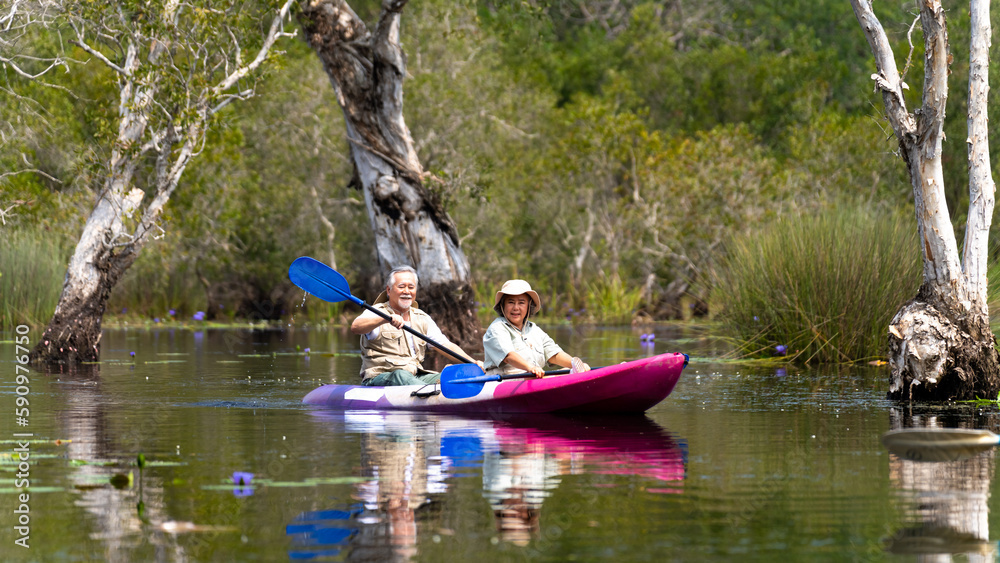 Asian senior couple kayaking together in the lake at mangrove forest on summer vacation. Retired elderly people man and woman have fun outdoor lifestyle travel nature and rowing a boat in the river.